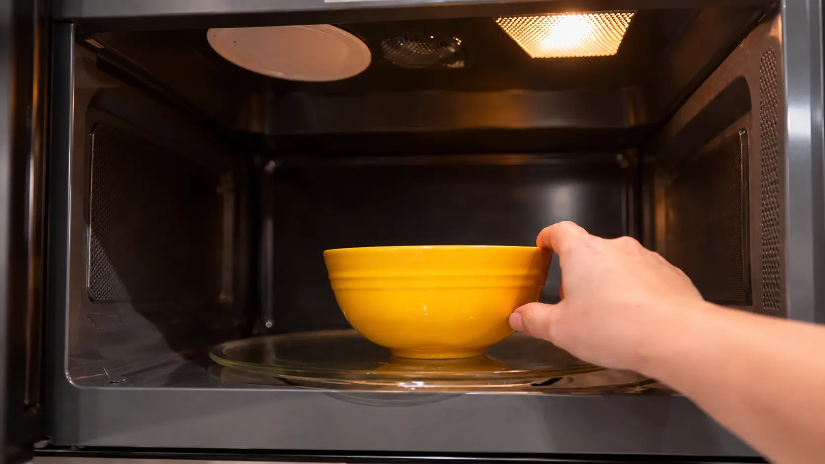 A hand places a bright yellow ceramic bowl onto the glass turntable inside a microwave. The microwave interior is warmly lit, highlighting the bowl against the dark stainless-steel interior, creating a clean, modern kitchen scene. The image captures the step of heating a pumpkin cheesecake mixture before chilling, with a professional food-photography style and soft depth of field.