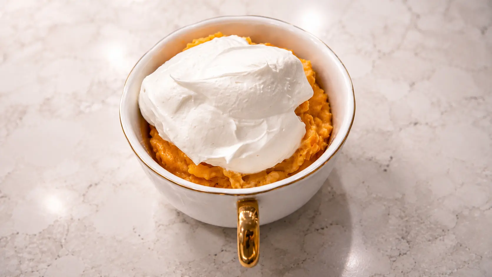 Overhead food photography of a no-bake pumpkin cheesecake served in a white ceramic cup with a gold handle on a light marble countertop. The creamy orange pumpkin cheesecake base is topped with a generous dollop of fluffy whipped topping, creating a smooth contrast between the vibrant pumpkin filling and the silky white cream. Soft natural lighting and a clean, minimalist composition give the dessert an elegant, professional food-photography appearance.
