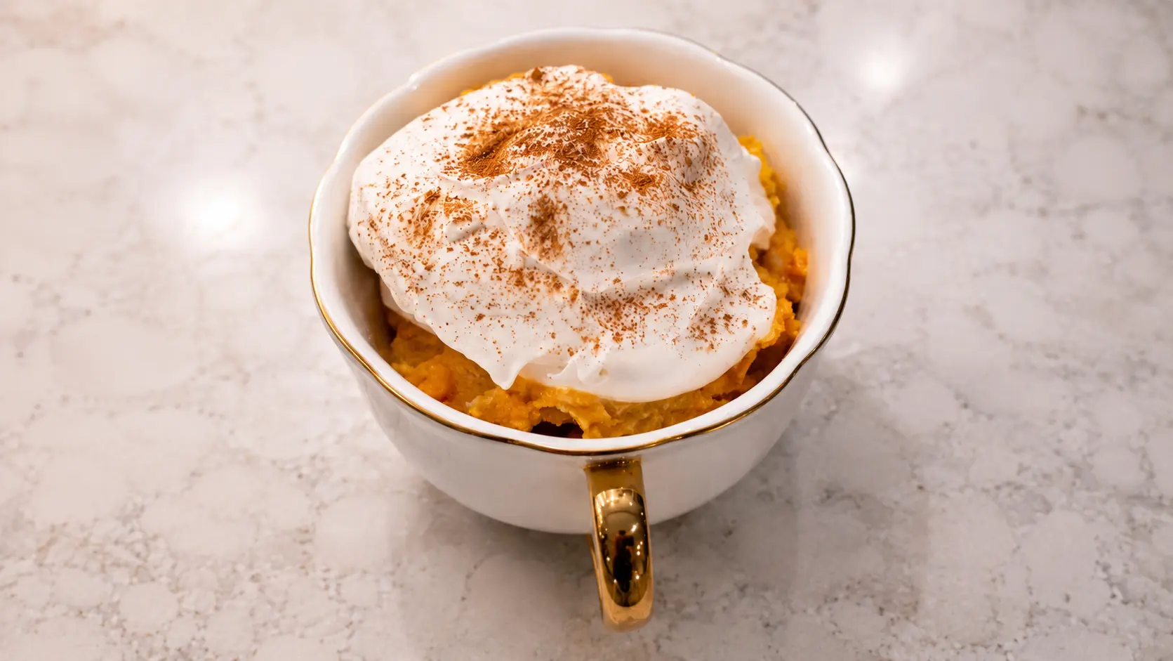 **Alt Text:** Close-up food photography of a no-bake pumpkin cheesecake served in a white scalloped ceramic cup with a gold handle. The creamy pumpkin cheesecake filling is topped with a thick layer of whipped topping and lightly dusted with cinnamon. Set on a light marble countertop, the dessert is photographed with soft natural lighting, highlighting the fluffy texture of the whipped cream and the rich orange pumpkin base for an elegant, bakery-style presentation.