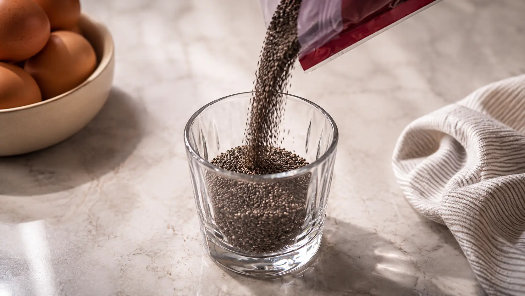 **Alt Text:** A professionally styled food photography image showing black chia seeds being poured from a package into a clear faceted glass on a light marble countertop. The falling seeds are captured in sharp detail, creating a dynamic sense of motion. Soft natural lighting highlights the texture of the chia seeds and the reflective glass surface, while a bowl of brown eggs and a neutral linen cloth sit blurred in the background, creating a warm, elegant kitchen atmosphere with a shallow depth of field.
