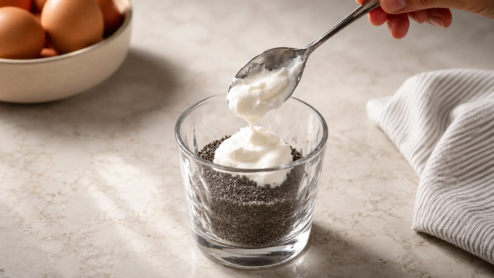 A professionally styled food photography image showing a spoonful of creamy Greek yogurt being added to a glass filled with black chia seeds on a light marble countertop. The yogurt slowly drips from the spoon, creating a sense of motion and freshness. Soft natural lighting highlights the smooth texture of the yogurt and the glossy glass, while a bowl of brown eggs and a neutral striped linen cloth appear softly blurred in the background. The composition uses shallow depth of field and warm tones to create an elegant, high-end recipe preparation scene.