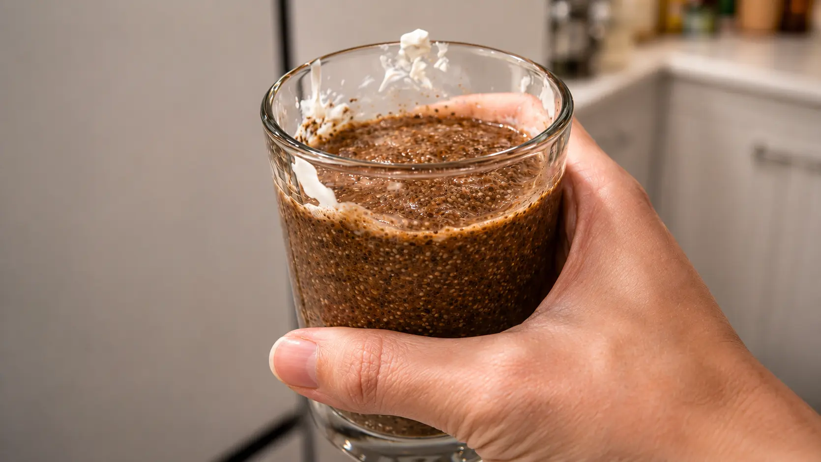 **Alt Text:** A professionally styled food photography image showing a hand holding a glass of freshly mixed tiramisu chia pudding in a bright modern kitchen. The pudding has a rich coffee-brown color with visible chia seeds suspended throughout, while traces of creamy yogurt cling to the inside rim of the glass. Soft natural lighting highlights the thick, velvety texture of the mixture, and the blurred kitchen background creates a clean, elegant atmosphere. The image captures the final preparation stage before refrigeration, with a warm, inviting, café-inspired aesthetic.