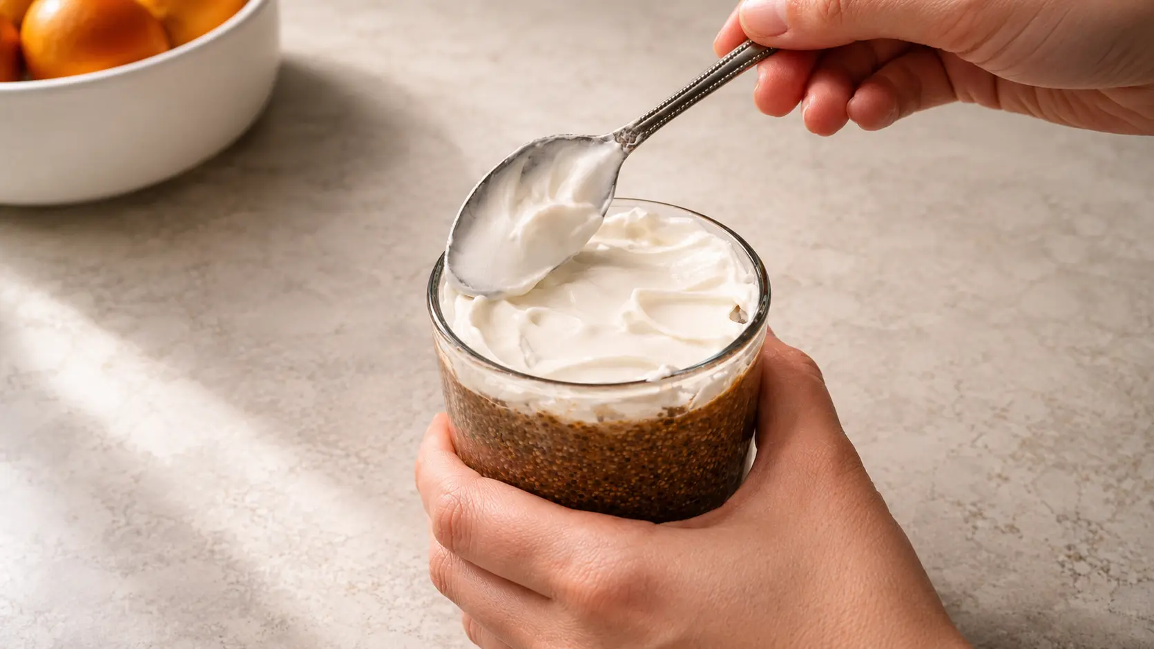 **Alt Text:** A professionally styled food photography image showing hands holding a glass of tiramisu chia pudding while spreading a thick layer of fluffy Cool Whip across the top with a spoon. The dessert features a rich coffee-colored chia pudding base visible through the clear glass, topped with a smooth, creamy white layer with elegant swirls. Soft natural lighting highlights the contrast between the dark pudding and airy topping, while a blurred bowl of fruit in the background and a neutral stone surface create a warm, upscale café-inspired atmosphere.