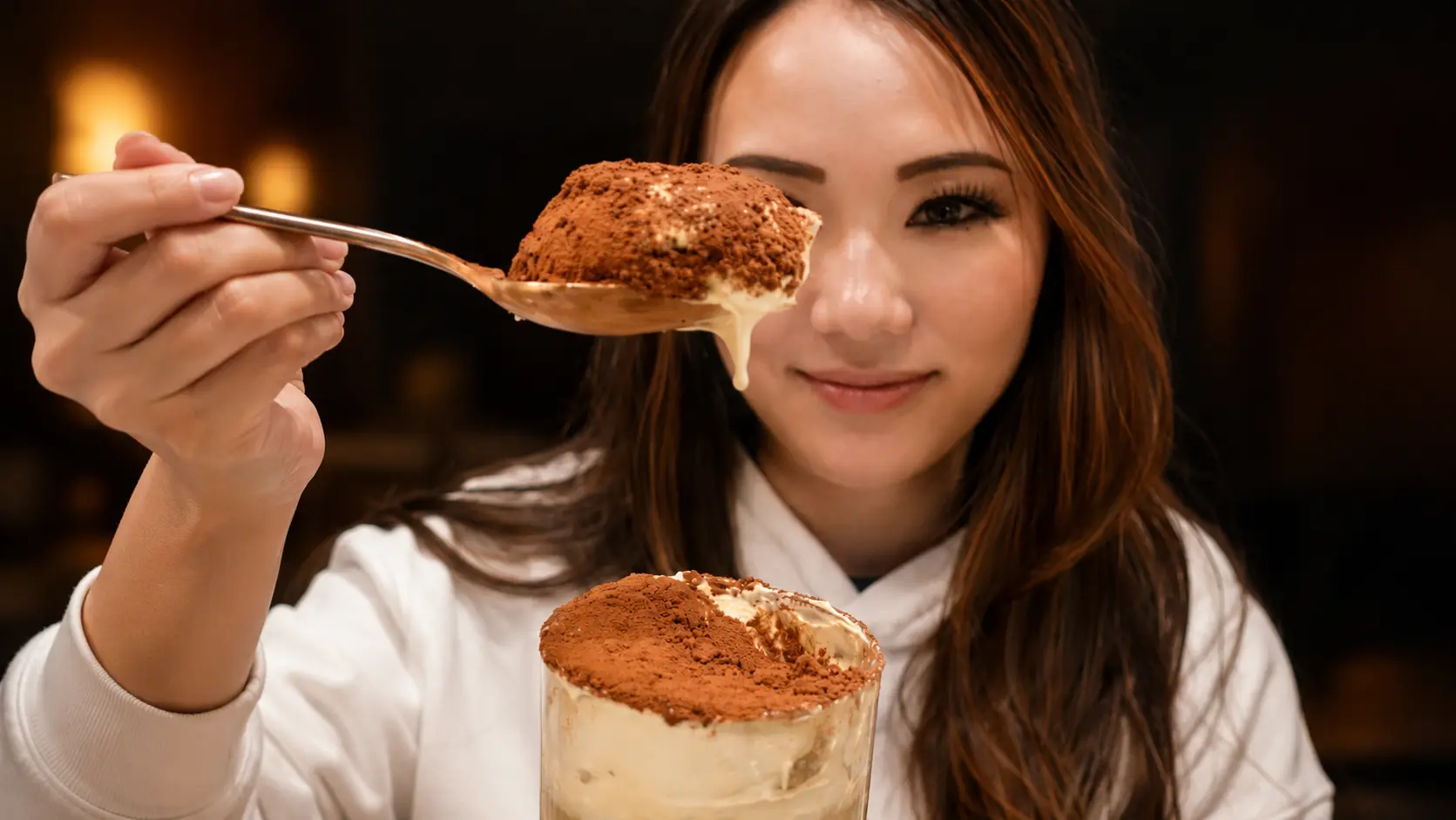 **Alt Text:** A professionally styled food photography portrait featuring a smiling woman holding a glass of tiramisu chia pudding topped with a thick layer of creamy whipped topping and a generous dusting of cocoa powder. In the foreground, a spoon lifts a rich, velvety bite of the dessert toward the camera, showcasing the fluffy cream and cocoa-coated surface with a delicate drip of cream. The layered chia pudding is visible through the clear glass, revealing its smooth, coffee-infused texture. Warm ambient lighting and a softly blurred background create an inviting café-style atmosphere that emphasizes both the dessert and the joyful tasting experience.