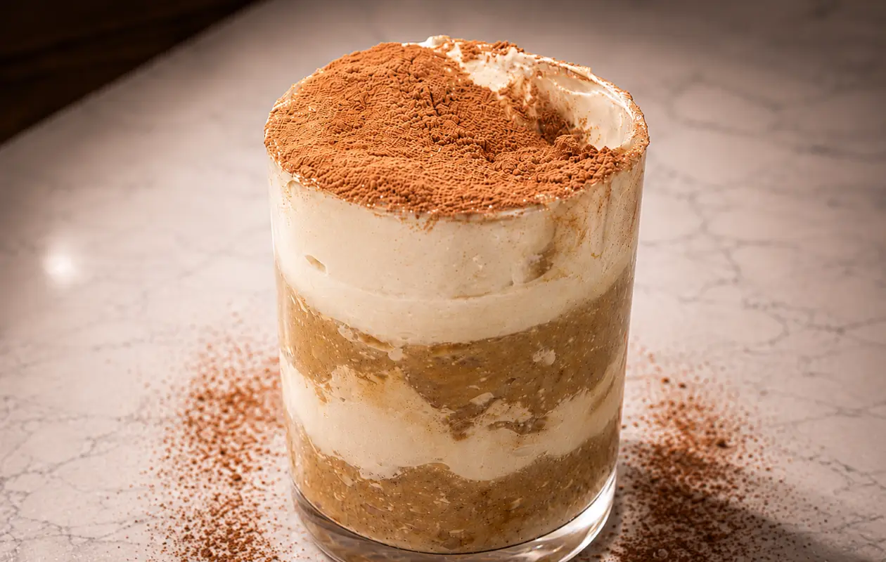 A professionally photographed tiramisu chia pudding parfait served in a clear glass cup on a light marble surface. Distinct layers of creamy mascarpone-style filling and coffee-infused chia pudding are visible through the glass, topped with a generous dust