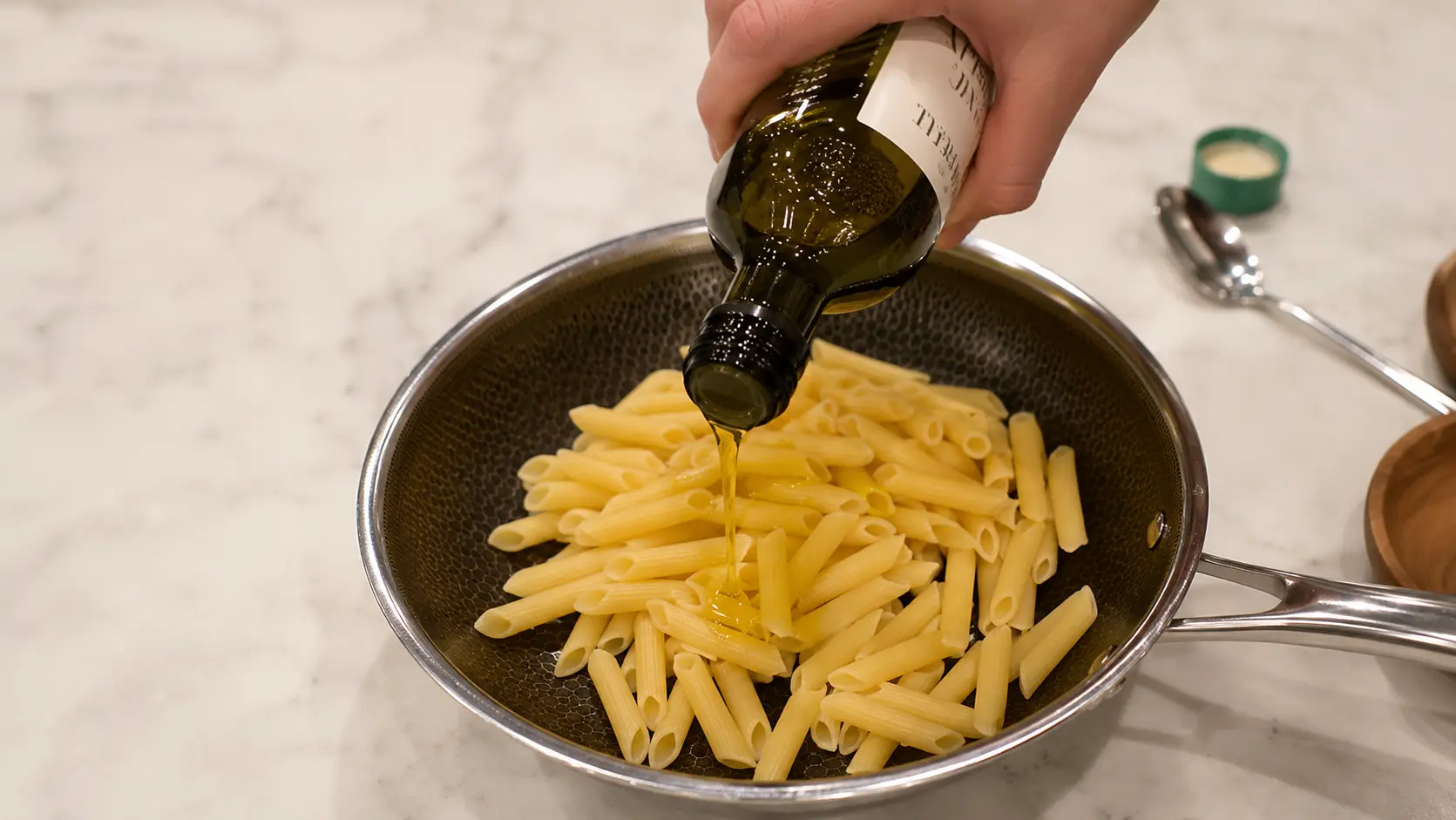 **Alt Text:**  *A professional food-photography image showing a hand drizzling golden olive oil from a dark glass bottle over cooked penne pasta in a stainless-steel skillet. The stream of olive oil is captured mid-pour, glistening under soft natural light. The pasta sits on a light marble countertop with subtle kitchen props blurred in the background, creating a clean, elegant cooking scene with sharp detail and warm, inviting tones.*