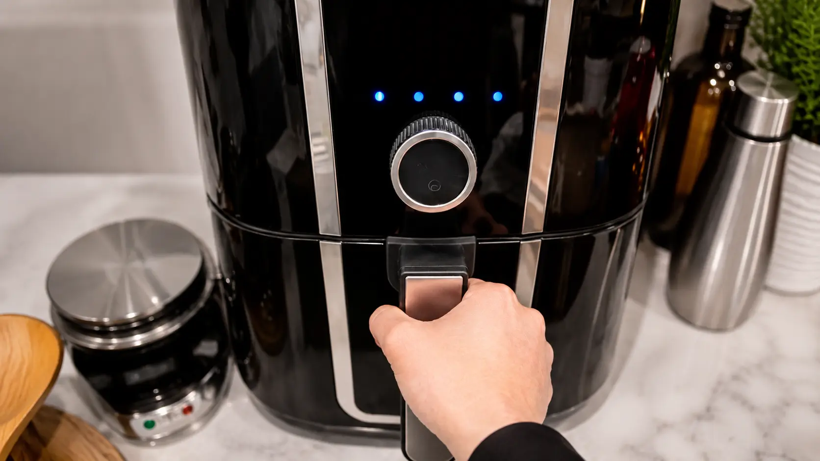 **Alt Text:**  *A professional food-photography image of a hand holding the handle of a glossy black air fryer on a light marble countertop. The air fryer’s blue indicator lights glow softly, suggesting it is actively cooking. The scene is brightly lit with clean reflections on the appliance’s polished surface, while blurred kitchen accessories in the background create depth and a modern home-kitchen atmosphere. The composition conveys the step of air-frying pasta chips to achieve a crispy, golden texture.*