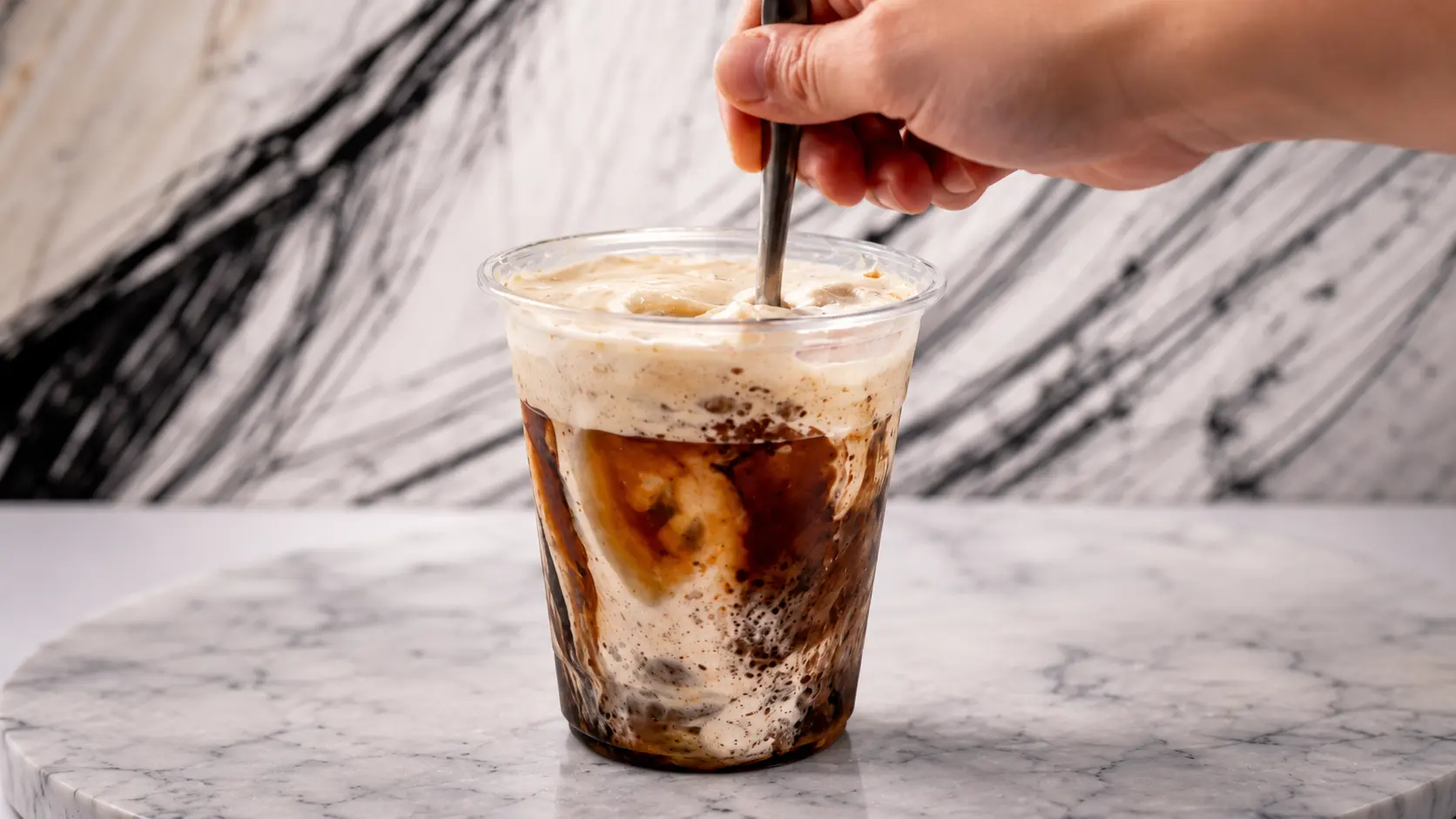 **Alt Text:** *A professional food-photography image of a creamy iced coffee dessert in a clear plastic cup resting on a white marble surface. Rich coffee swirls blend with a thick, velvety cream mixture, creating a dramatic marbled effect throughout the drink. A hand stirs the dessert with a spoon from above, capturing the moment the coffee and cream are mixing together. Soft studio lighting highlights the glossy textures, while the elegant marble backdrop and shallow depth of field create a modern café-style presentation.*