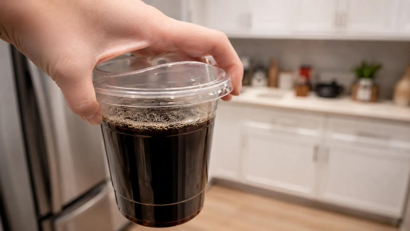 **Alt Text:** *A professional food-photography image of a hand holding a clear cup filled with freshly brewed black coffee in a bright modern kitchen. Tiny bubbles line the surface of the coffee, highlighting its freshly brewed appearance. The rich, dark coffee contrasts beautifully with the transparent cup, while the softly blurred kitchen cabinets and stainless-steel refrigerator in the background create depth and a clean, contemporary atmosphere. Warm natural lighting gives the scene a cozy, inviting feel, capturing the step before the coffee mixture is chilled to make Japanese coffee jelly.*