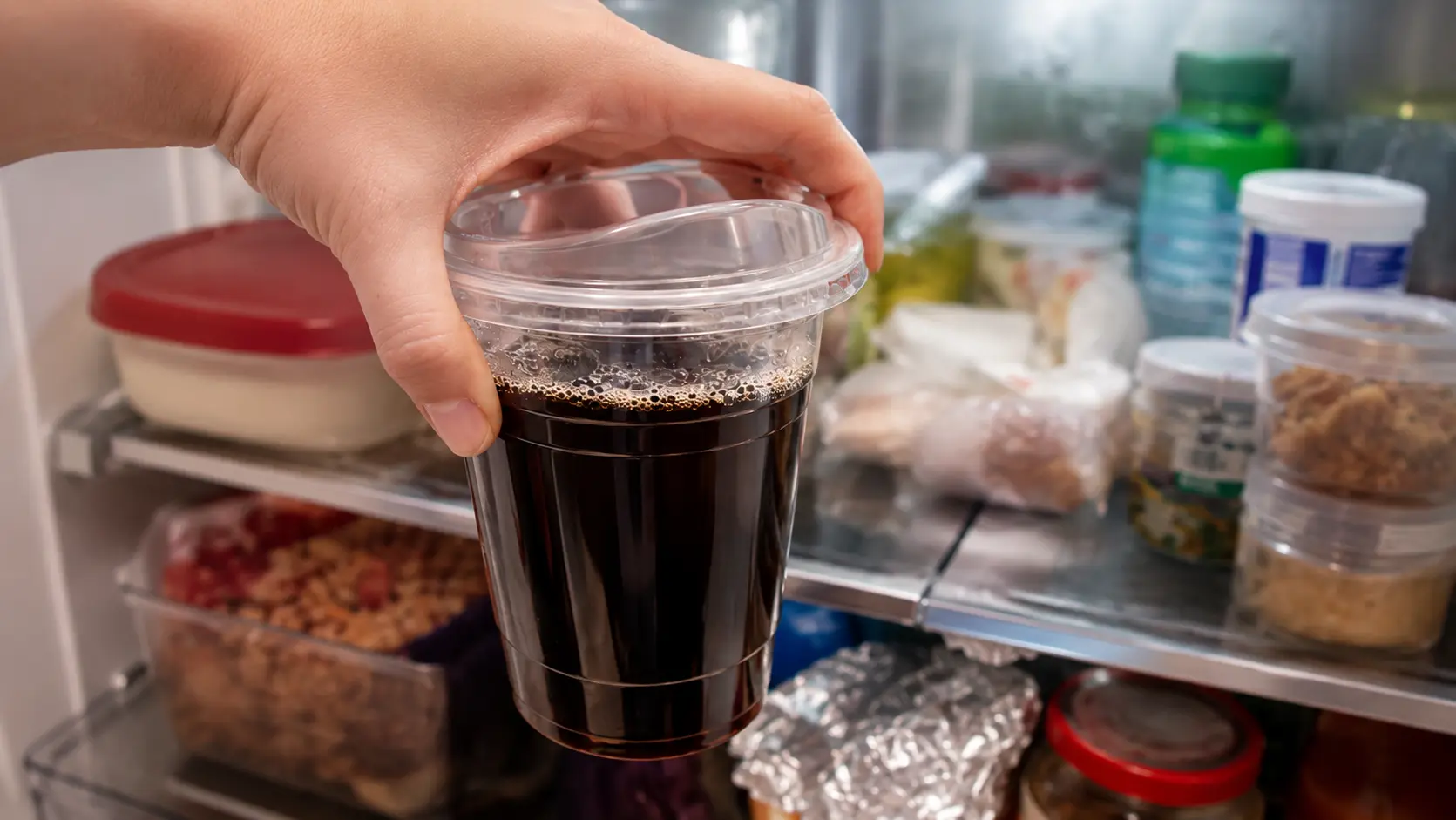 **Alt Text:** *A professional food-photography image of a hand placing a clear plastic cup filled with dark coffee into an open refrigerator. The coffee appears rich and glossy, with a thin ring of tiny bubbles along the surface. The refrigerator interior is softly blurred, showing neatly stored containers and fresh ingredients in the background. Bright, cool lighting emphasizes the contrast between the deep black coffee and the clean refrigerator environment, capturing the step of chilling the coffee mixture so it can set into Japanese coffee jelly.*