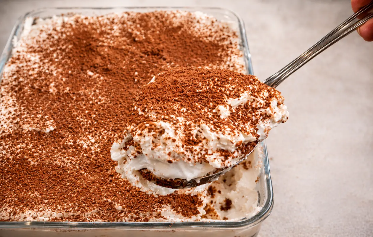 **Alt text:** A close-up of a creamy homemade tiramisu in a clear glass baking dish, topped with a light dusting of cocoa powder. A spoon lifts a generous serving, revealing fluffy mascarpone layers and coffee-soaked cake beneath. The dessert is photograph