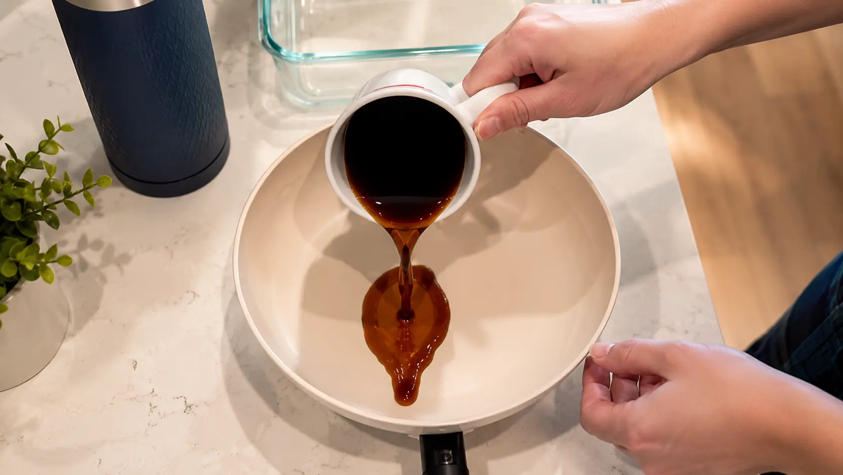 Professional food photography overhead shot of freshly brewed coffee being poured from a white ceramic mug into a shallow cream-colored dish on a light marble countertop. A clear glass baking dish and insulated bottle sit nearby, creating a clean kitchen prep scene. Warm natural light highlights the rich dark coffee and soft neutral tones, giving the image a polished, editorial-style cooking aesthetic.