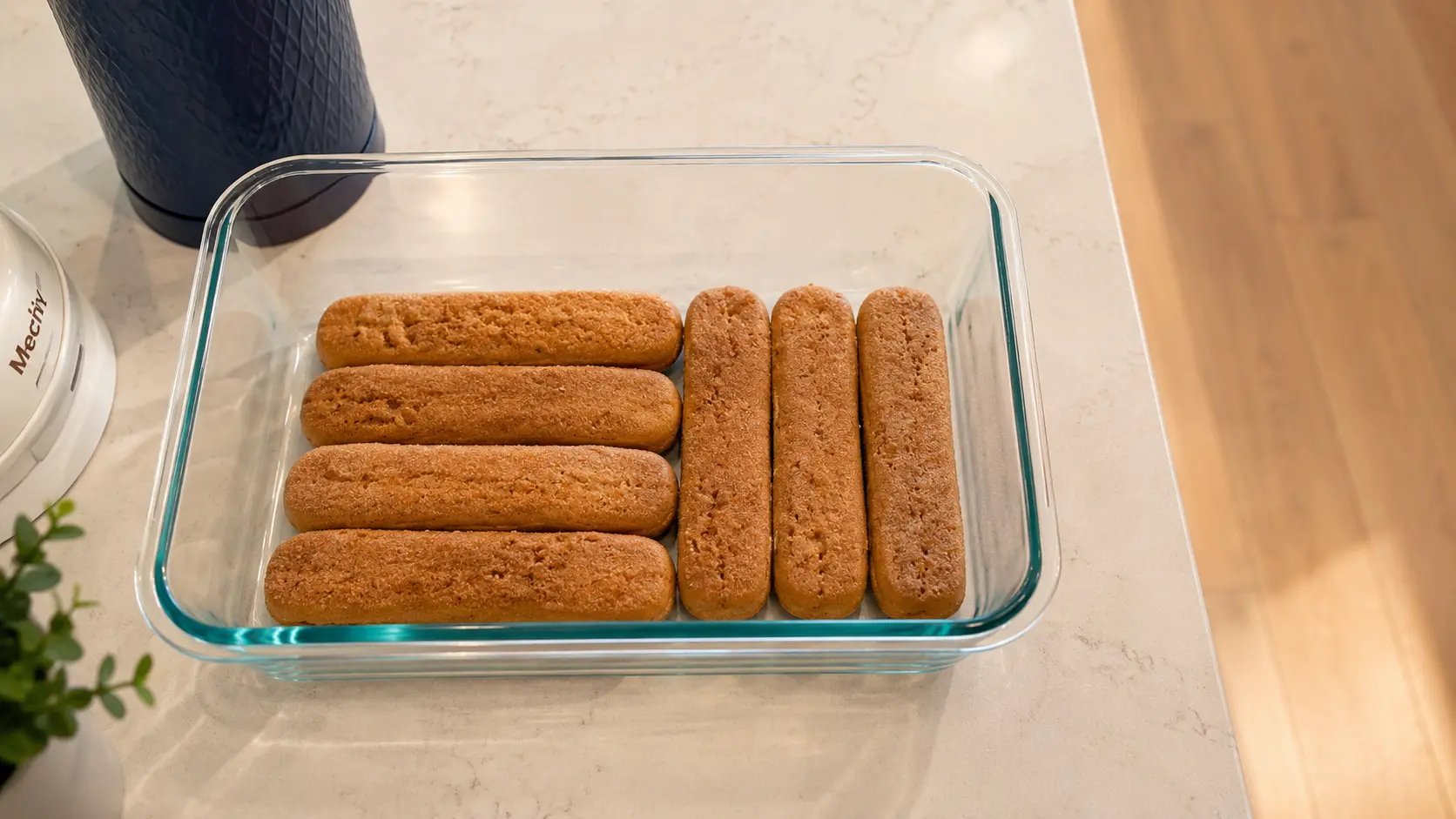 Professional food photography overhead shot of a rectangular glass baking dish filled with a neatly arranged layer of coffee-soaked ladyfinger cookies for tiramisu. The dish sits on a light marble countertop with soft natural lighting that highlights the golden-brown texture of the cookies and the clean glass edges. The bright, airy kitchen setting creates a polished, inviting dessert-preparation scene.