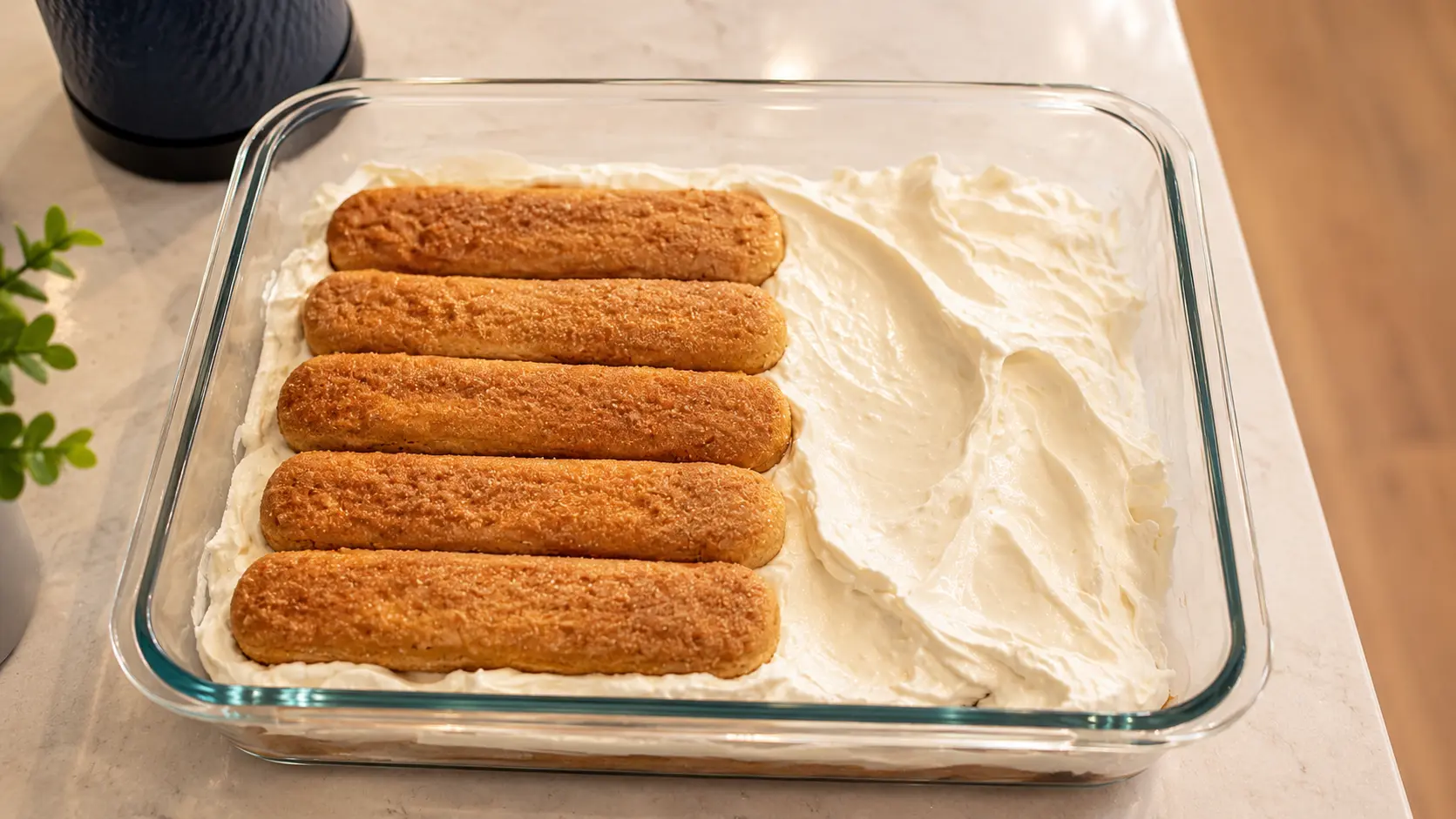 Professional food photography close-up of a partially assembled tiramisu in a clear glass baking dish. A smooth layer of mascarpone cream is spread across coffee-soaked ladyfinger cookies, while a second layer of golden-brown ladyfingers is neatly arranged on top. The dessert sits on a light marble countertop with soft natural lighting that highlights the creamy texture, crisp cookie details, and clean glass edges, creating an elegant, bakery-quality presentation.