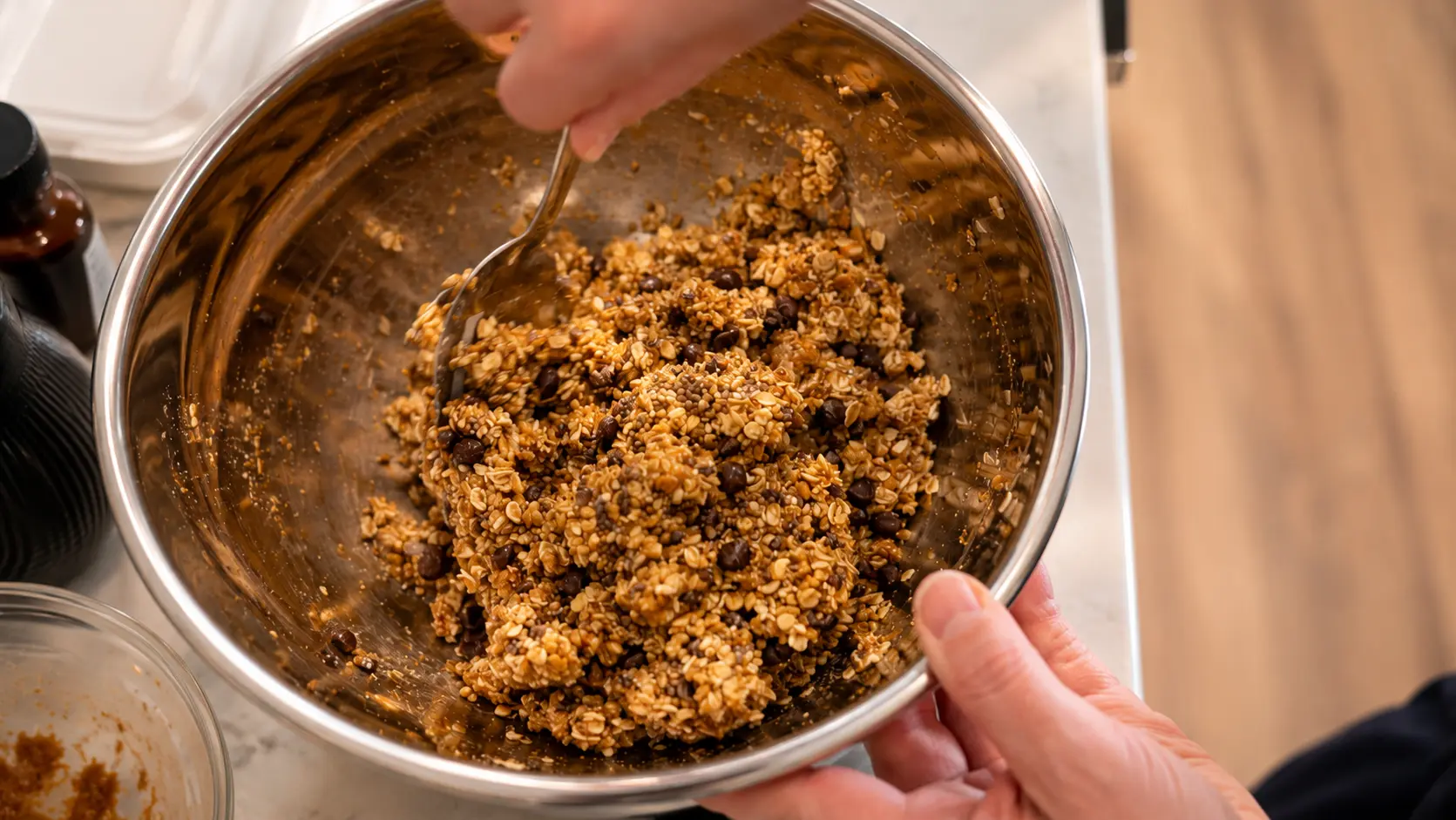 Professional overhead food photograph of a homemade energy ball mixture being stirred in a large stainless steel mixing bowl. The mixture contains rolled oats, chocolate chips, chia seeds, peanut butter, honey, and cinnamon, creating a chunky, golden-brown texture. One hand holds the bowl steady while another stirs with a spoon. Warm natural lighting highlights the ingredients, and the softly blurred marble countertop and kitchen items create a clean, inviting, and high-end recipe-preparation atmosphere.