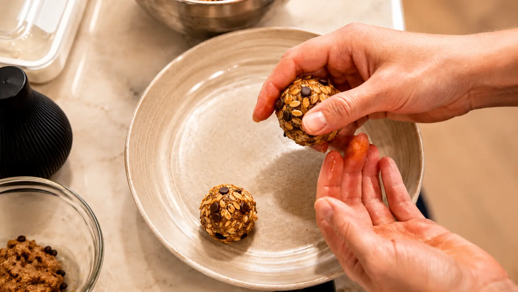 Professional food photograph showing hands rolling homemade energy balls made from oats, chocolate chips, chia seeds, peanut butter, honey, and cinnamon. One finished energy ball rests on a light beige ceramic plate while another is being shaped by hand above it. Warm natural lighting highlights the glossy texture of the mixture and the visible oats and chocolate chips. The softly blurred countertop, mixing bowl, and preparation ingredients in the background create a clean, inviting, and high-end recipe-making atmosphere.