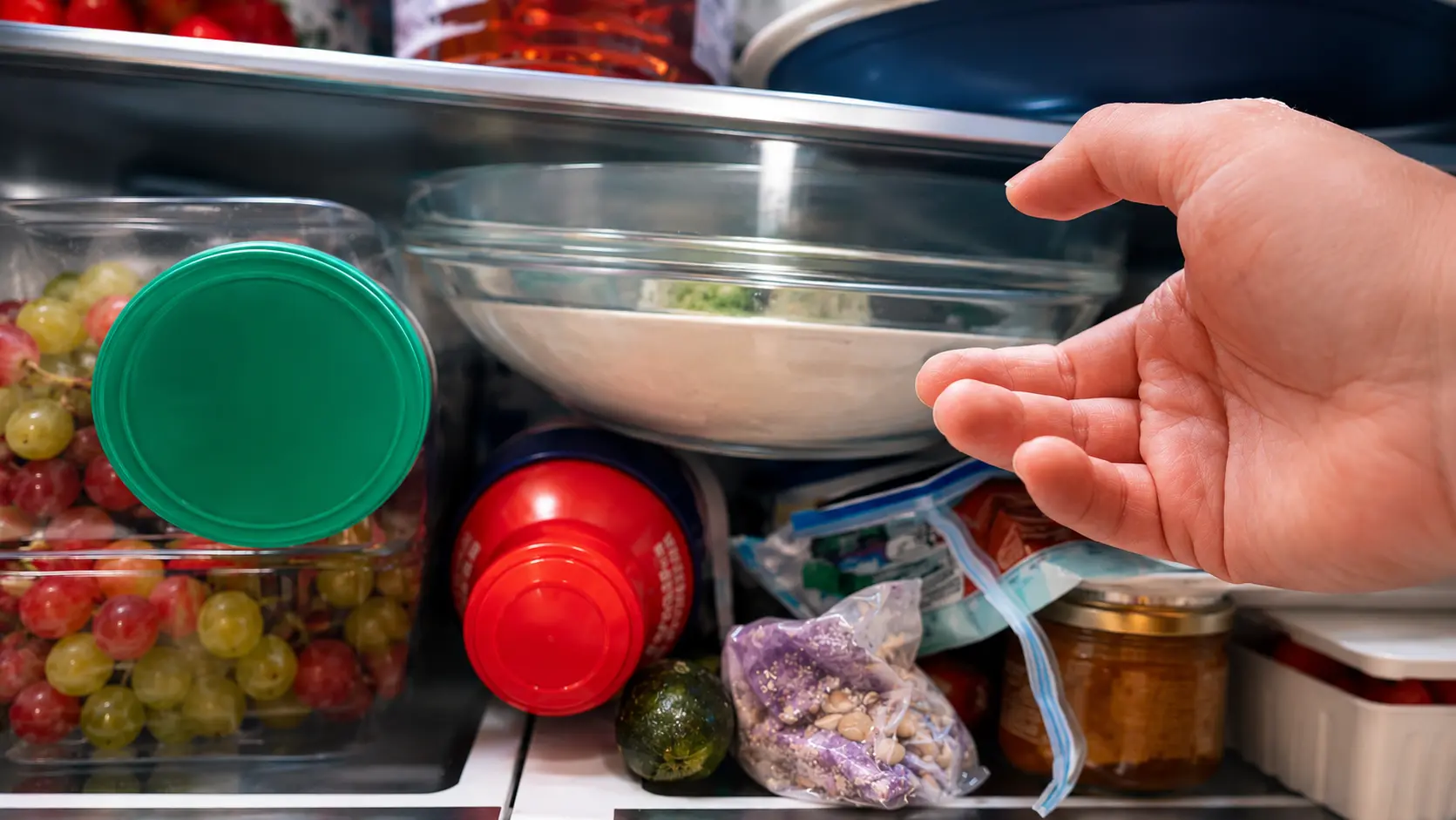 Hand reaching into a refrigerator toward a glass bowl of chilled mixture, surrounded by fresh grapes, produce, and stored ingredients on neatly organized shelves. The image is brightly lit with crisp detail, creating a clean, professional food-photography style that emphasizes freshness and meal preparation.