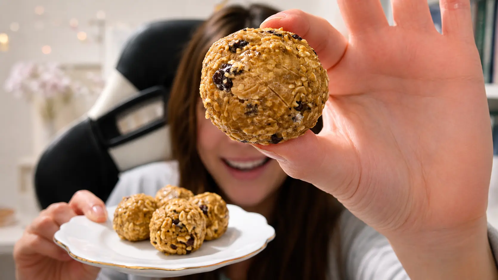 Close-up of a homemade oatmeal energy bite held toward the camera, showcasing its chewy texture with oats, seeds, and chocolate chips. In the softly blurred background, a smiling woman holds a white scalloped plate filled with additional energy bites, creating a warm, inviting food-photography scene with natural lighting and shallow depth of field.