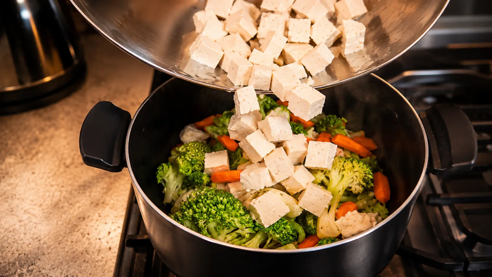 A professional food photography shot capturing cubed tofu being poured from a stainless steel mixing bowl into a black cooking pot filled with vibrant broccoli, carrots, and cauliflower on a stovetop. The fresh vegetables appear bright and crisp, while wisps of steam rise from the pot, creating a warm, inviting kitchen atmosphere. Soft natural lighting highlights the textures of the tofu and vegetables, with a shallow depth of field gently blurring the background for a polished, restaurant-quality presentation.