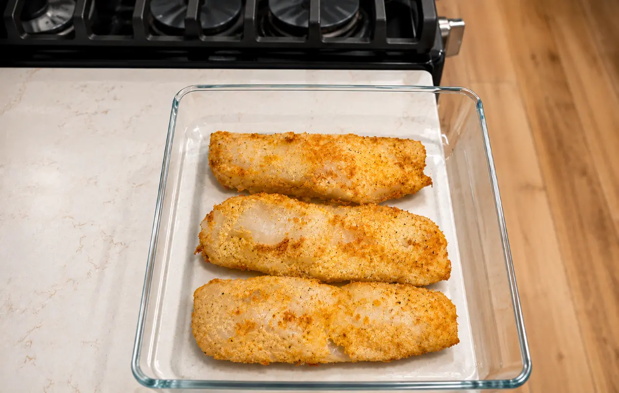 Golden-brown breaded fish fillets arranged in a clear glass baking dish on a light marble countertop, photographed from above in a bright modern kitchen. The crispy coating and tender white fish are highlighted with soft natural lighting, creating a clean,