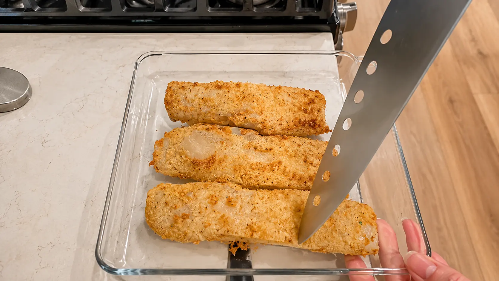 Overhead food photography of three crispy, golden-breaded fish fillets arranged in a clear glass baking dish on a light marble countertop. A stainless-steel spatula slides underneath one fillet while a hand steadies the dish. Warm kitchen lighting highlights the crunchy texture and golden coating, with a modern stovetop and wood flooring softly blurred in the background.