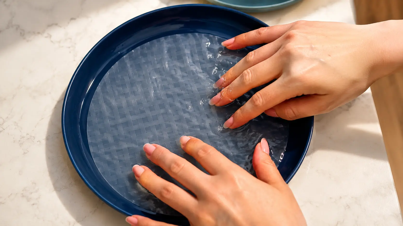 Close-up overhead food preparation photo showing two hands gently softening a translucent rice paper sheet in a deep navy-blue bowl filled with water. The bowl sits on a light marble countertop, illuminated by soft natural light that highlights the delicate woven texture of the rice paper. The clean, minimalist composition creates an elegant, professional food photography aesthetic ideal for a recipe tutorial.