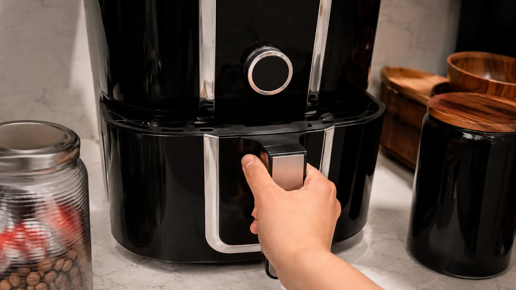 Professional food photography featuring a sleek black air fryer on a clean marble countertop. A hand grips the air fryer basket handle, preparing to slide it open, while stylish kitchen accessories and storage containers sit softly blurred in the background. Warm natural lighting highlights the glossy finish of the appliance, creating an inviting step in a homemade mozzarella stick recipe tutorial.