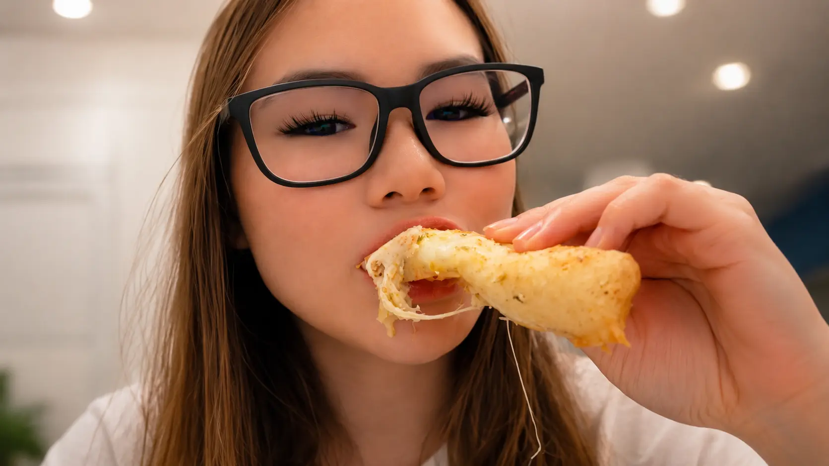 Close-up food photography of a woman wearing black-framed glasses taking a bite of a crispy rice paper mozzarella stick. Gooey melted mozzarella stretches from the golden-brown snack as she holds it near her mouth, with warm indoor lighting and a softly blurred kitchen background creating a cozy, appetizing atmosphere. The focus highlights the crispy exterior and cheesy pull, emphasizing the snack’s irresistible texture.