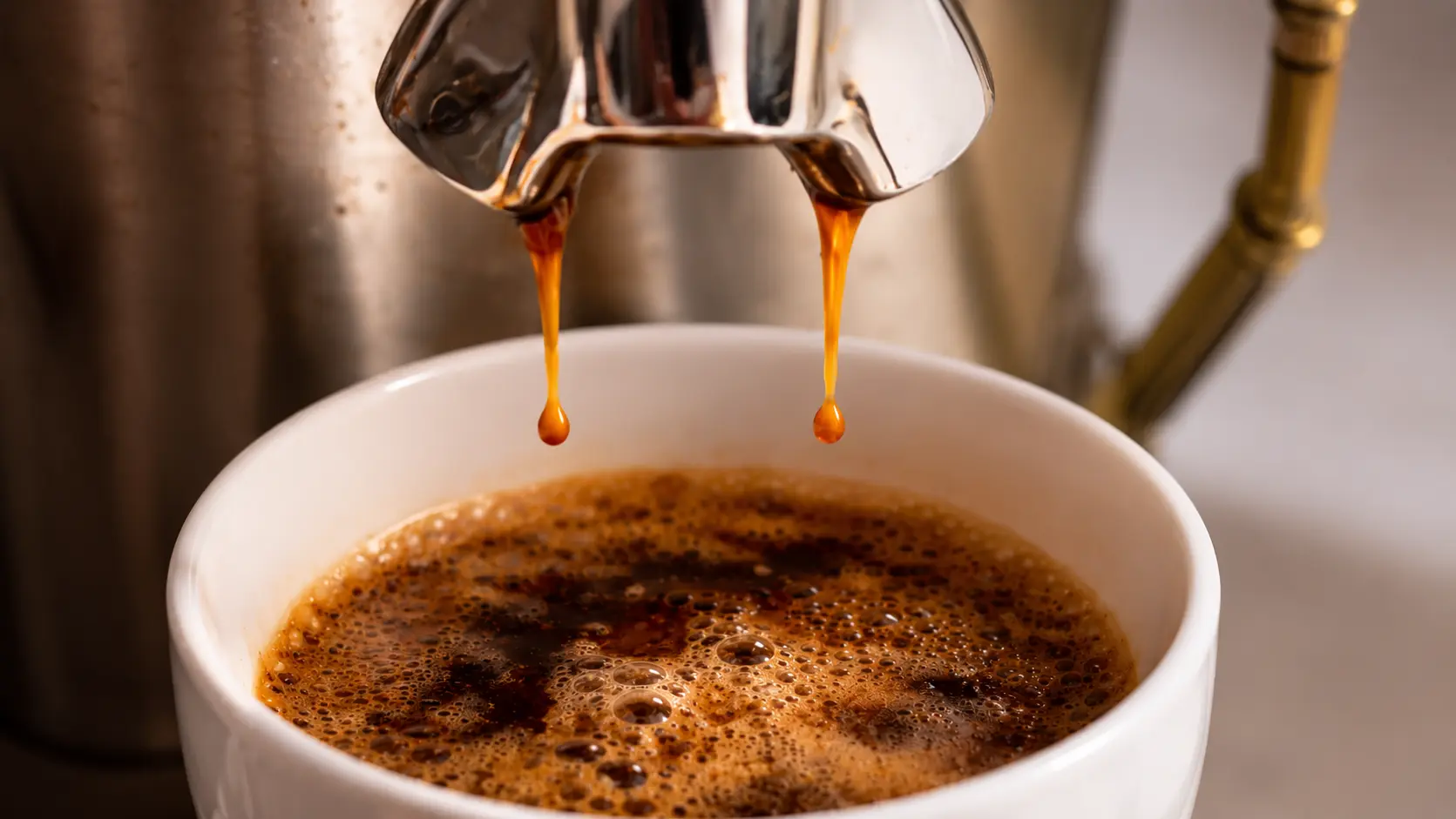 Professional food photography close-up of rich espresso dripping from a polished stainless steel portafilter into a white ceramic cup. Two golden-brown streams of freshly extracted espresso fall into a layer of thick crema with glossy bubbles and swirling coffee patterns. Warm café lighting, shallow depth of field, and soft background blur highlight the texture and color of the espresso, creating an inviting artisan coffee scene.