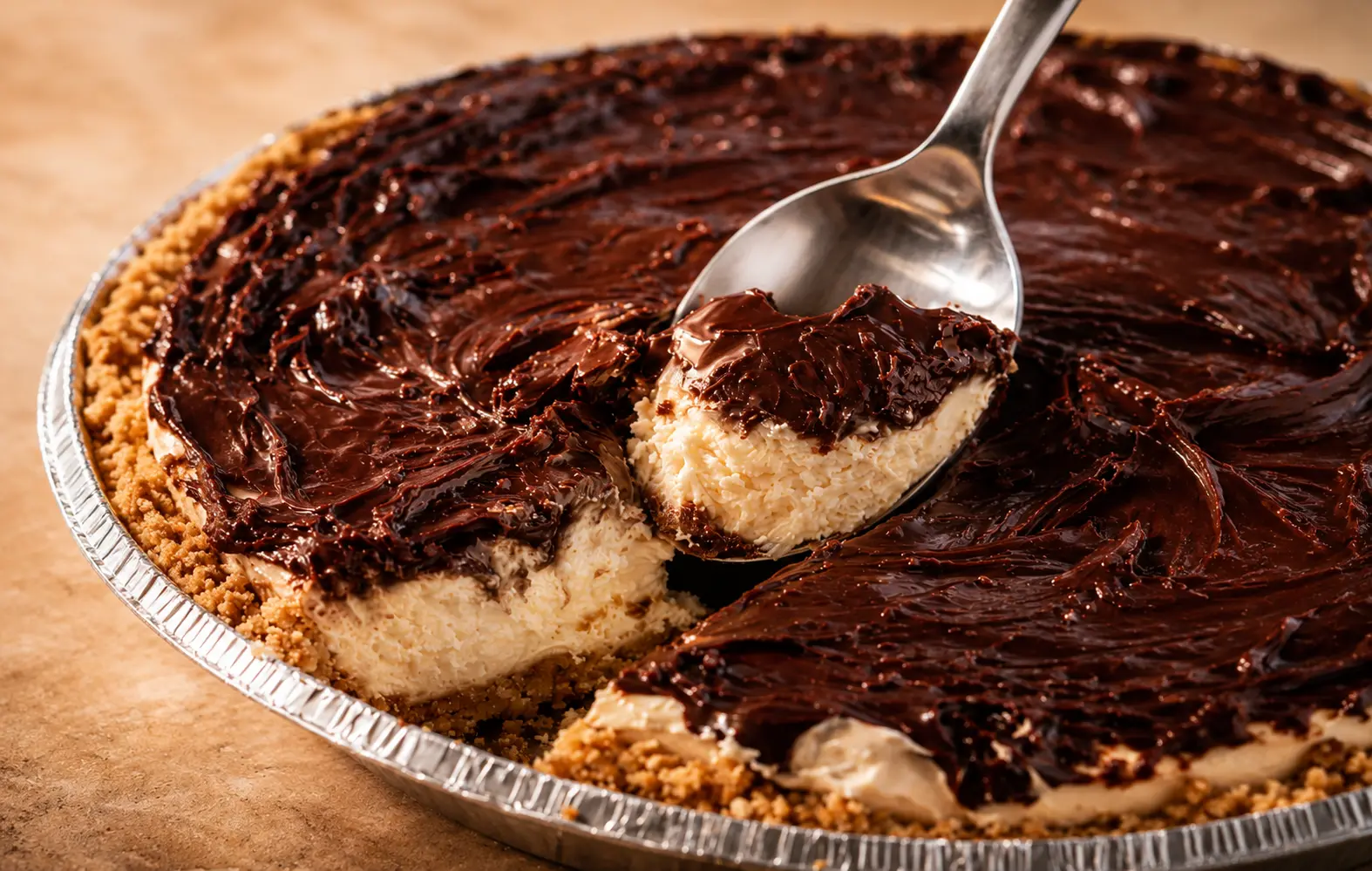 Close-up of a no-bake chocolate cheesecake with a buttery graham cracker crust and creamy vanilla filling, topped with a thick layer of glossy chocolate ganache. A spoon lifts a rich slice from the cheesecake, showcasing the smooth, fluffy texture under warm professional food photography lighting.