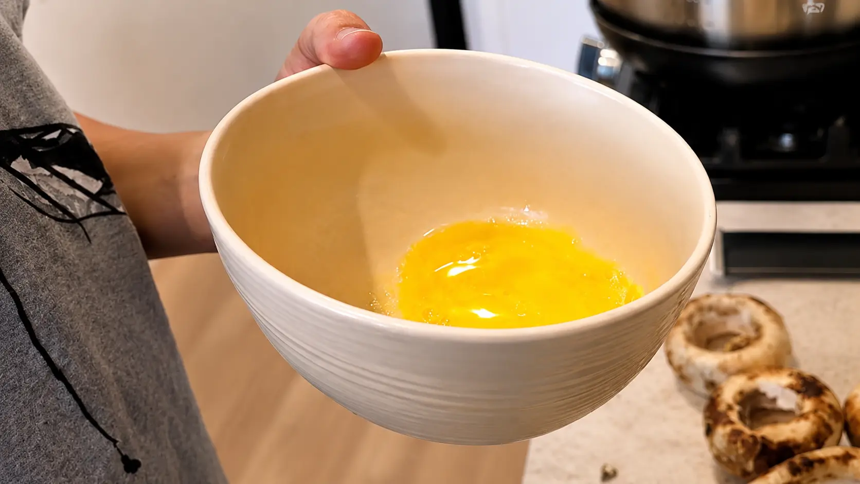 Professional food photography-style close-up of a person holding a large cream-colored ceramic mixing bowl containing melted butter in a bright home kitchen. Fresh mushroom caps rest on the countertop nearby, ready for stuffing. The image features warm natural lighting, realistic textures, soft background blur, and a clean culinary preparation setting that highlights the ingredients for a stuffed mushroom recipe.