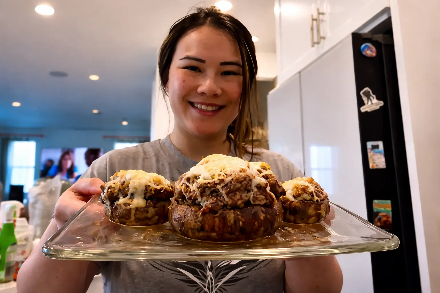 Smiling woman in a kitchen holding a glass plate of freshly baked stuffed mushrooms topped with melted cheese, presented close to the camera under warm lighting.