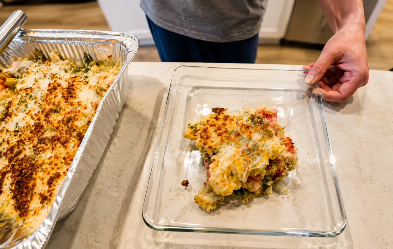 A professionally photographed baked vegetable casserole with a golden-brown cheese topping sits in a large aluminum baking tray on a bright kitchen countertop. A serving of the casserole is plated in a clear glass baking dish, showing layers of vegetables,