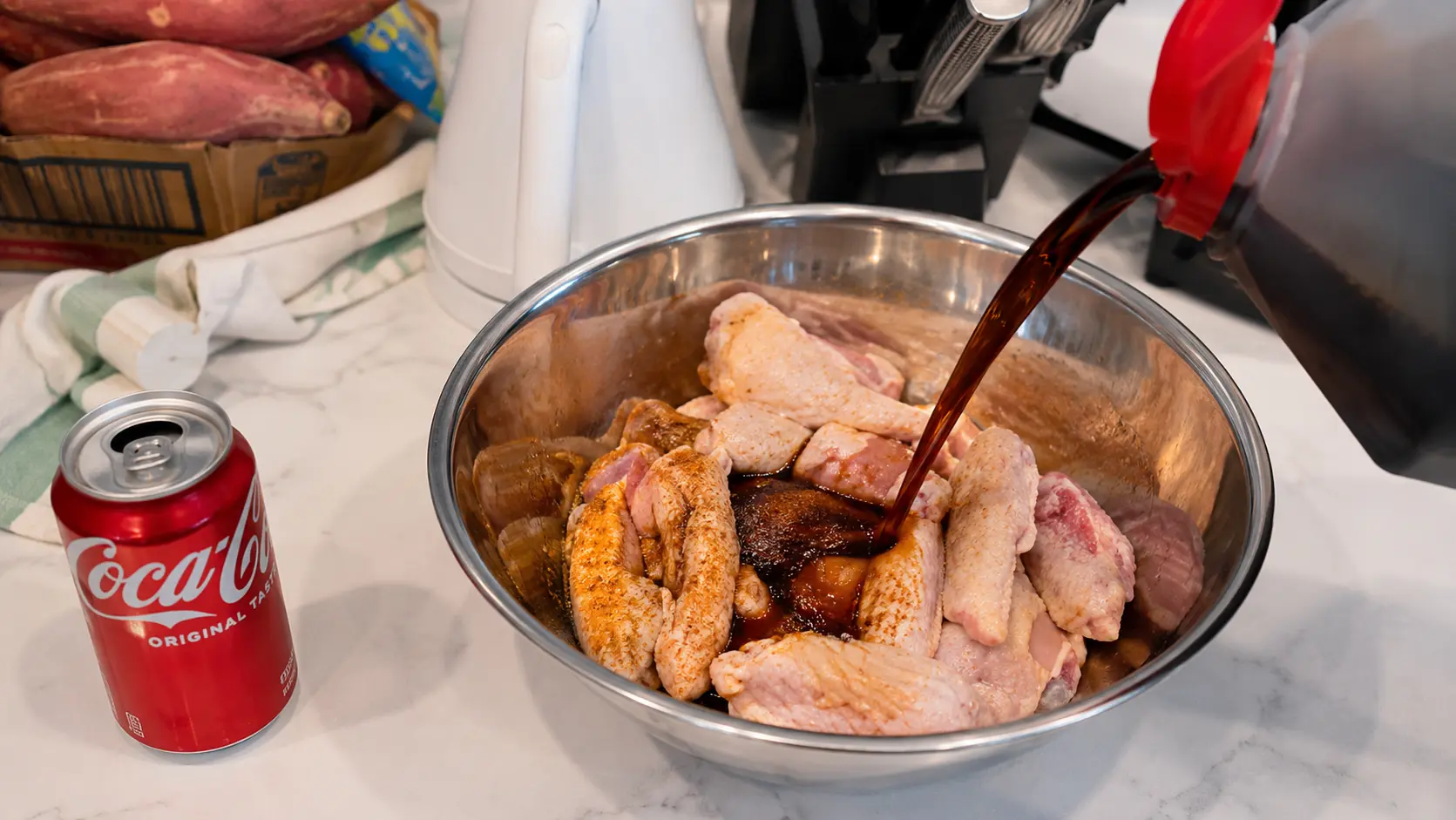 A professional food photography image showing raw chicken wings being marinated in a large stainless steel mixing bowl on a bright white marble countertop. A stream of dark soy sauce is being poured over the chicken from a plastic container with a red flip-top lid, capturing the action mid-pour. A red can of Coca Cola sits beside the bowl, hinting at the recipe's key ingredient. In the softly blurred background are sweet potatoes in a wooden crate, a white electric kettle, a knife block, and a folded kitchen towel. The lighting is bright and natural, highlighting the texture of the chicken, the glossy marinade, and the reflective surface of the metal bowl. The image has a clean, realistic, and inviting food blogger aesthetic, perfect for a Coca Cola chicken wings recipe tutorial.