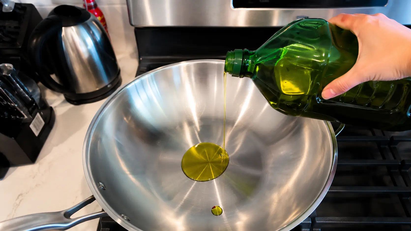 A professional food photography image showing olive oil being poured from a large green bottle into a shiny stainless steel wok on a stovetop. A golden stream of oil flows into the center of the pan, creating a reflective pool against the polished metal surface. The modern kitchen background includes a stainless steel kettle, knife block, and stove controls softly blurred to keep the focus on the cooking process. Bright natural lighting highlights the glossy olive oil, clean cookware, and sleek kitchen surfaces. The image has a realistic, high-end food blogger aesthetic, capturing the first step of preparing Coca Cola chicken wings with crisp detail and warm, inviting tones.