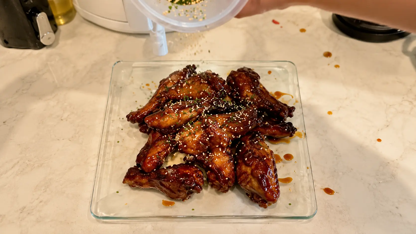 A professional food photography image showcasing a stack of Coca Cola chicken wings arranged on a flat transparent glass plate. The wings are coated in a thick, glossy caramelized Coca Cola and soy sauce glaze that reflects the warm kitchen lighting. A hand at the top of the frame sprinkles furikake seasoning over the wings, capturing the finishing garnish in midair. The glaze appears sticky and rich, with small droplets of sauce artistically scattered across the light marble countertop surrounding the plate. The transparent glass plate allows the countertop to show through, creating a clean and modern presentation. Softly blurred kitchen appliances in the background add depth while keeping the focus on the beautifully glazed wings. The image has a realistic, high-end food blogger aesthetic and captures the final step of preparing Coca Cola chicken wings before serving.