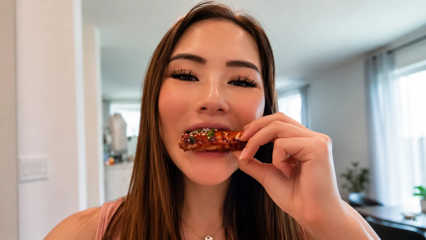 A professional lifestyle food photography image featuring a woman taking a bite of a glossy Coca Cola chicken wing. The wing is coated in a rich caramelized Coca Cola and soy sauce glaze and garnished with sesame seeds and furikake seasoning. The woman holds the wing near her mouth while smiling softly, showcasing the sticky, shiny texture of the glaze. Her long brown hair frames her face, and the background features a bright modern home interior with large windows and soft natural light. The shallow depth of field keeps the focus on the chicken wing and the tasting moment, creating an inviting food blogger aesthetic. The image captures the enjoyment of eating homemade Coca Cola chicken wings and conveys the delicious, restaurant-quality appeal of the recipe.