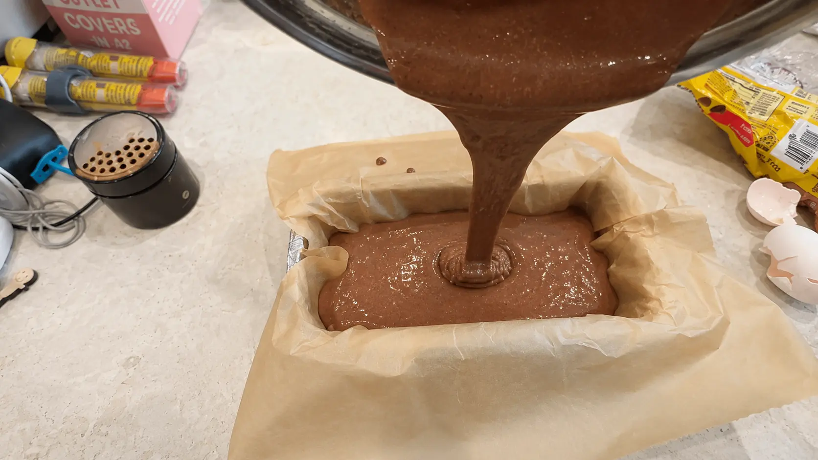 Pouring flourless high protein brownie batter into a parchment lined baking pan. The rich chocolate mixture is made with banana, eggs, Greek yogurt, honey, and cocoa powder.