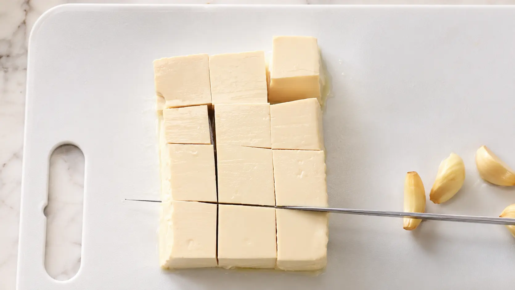 Fresh tofu being cut into cubes on a white cutting board with garlic cloves, ready for cheesy Shin ramen noodles.