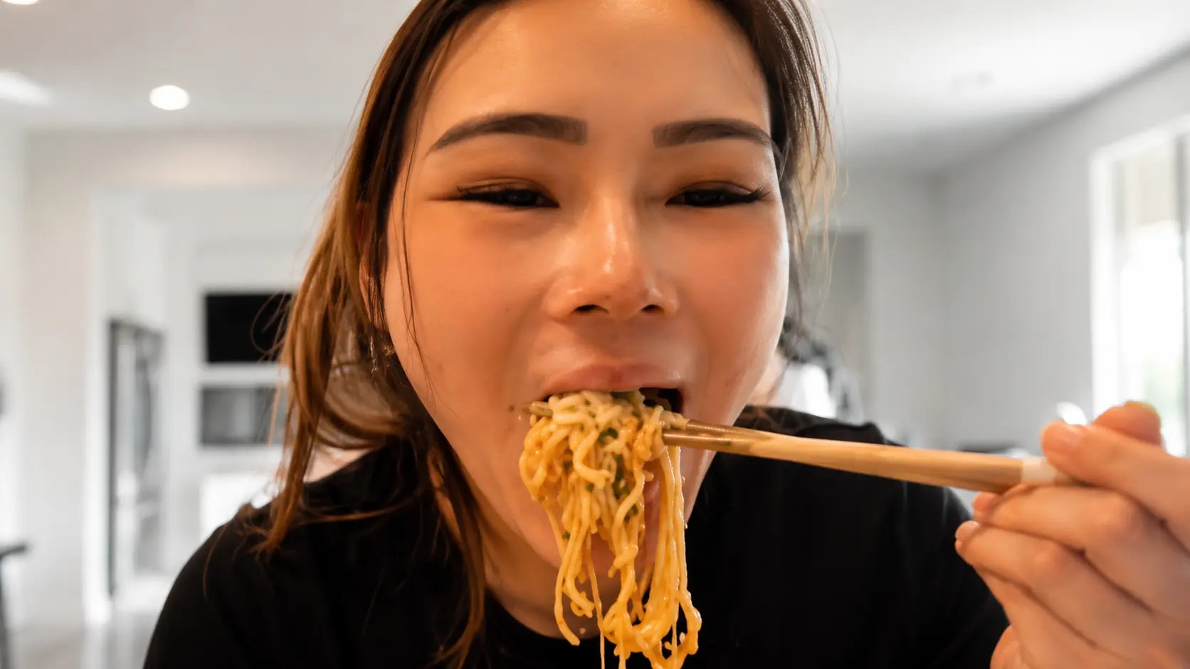 Woman enjoying cheesy ramen noodles with chopsticks in a bright modern kitchen.