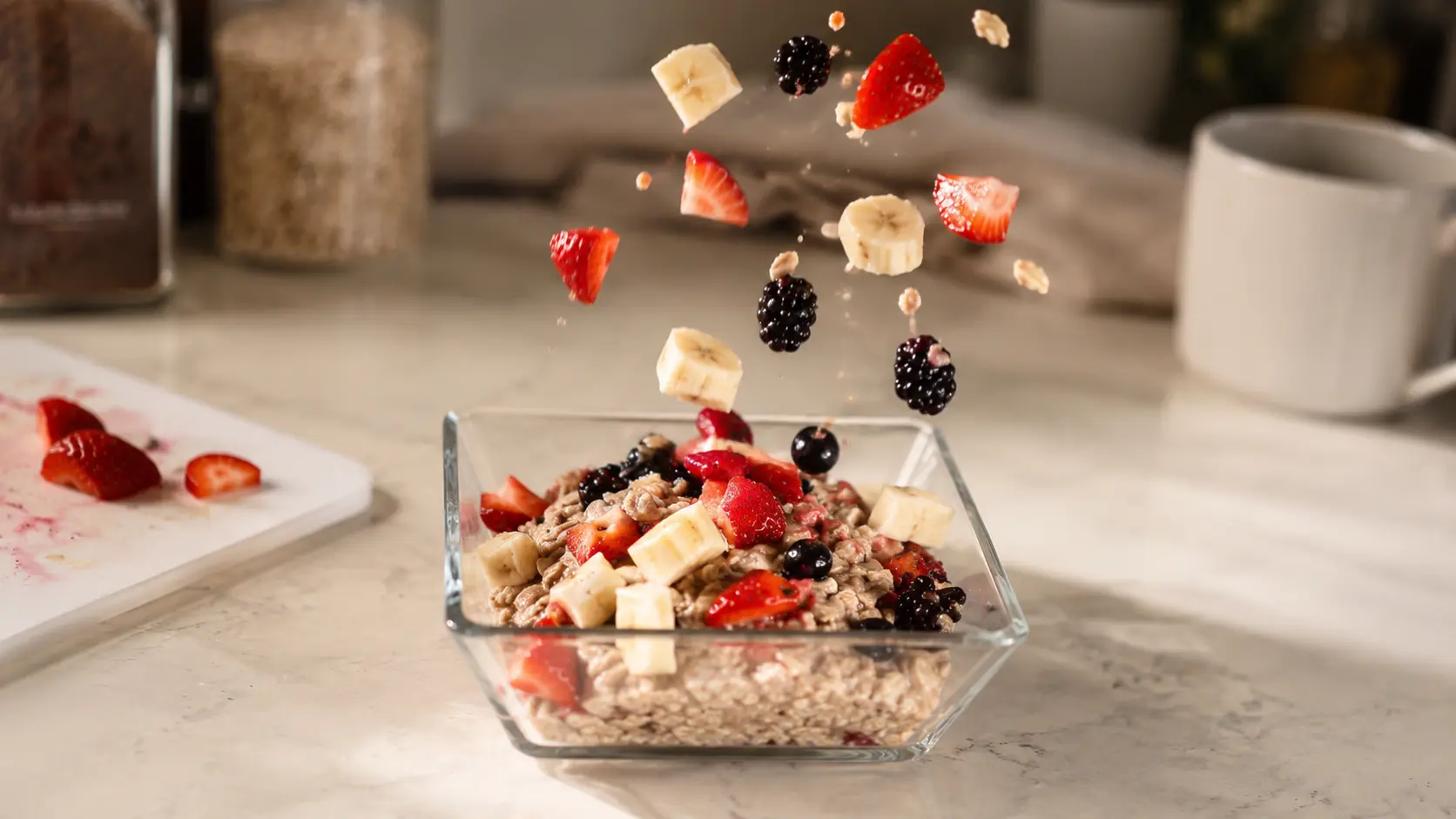 fruits like banana, strawberries, and blackberries falling onto a glass bowl of oatmeal