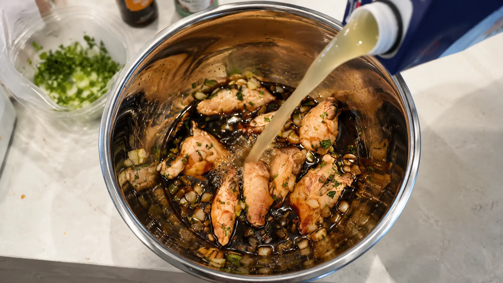 Chicken broth being poured into a bowl of marinated chicken wings with onion, garlic, and cilantro.