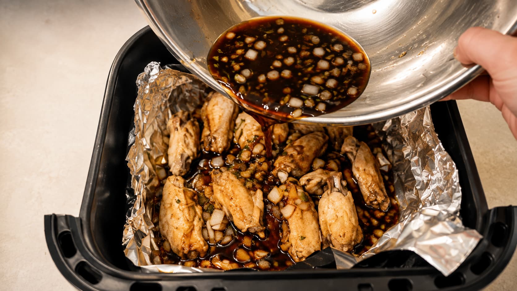Marinade with onion and garlic being poured over chicken wings in a foil-lined air fryer basket.
