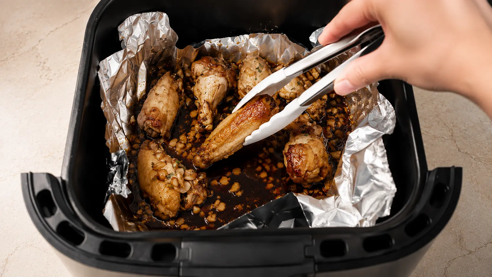 Using tongs to flip chicken wings in a foil-lined air fryer basket halfway through cooking.