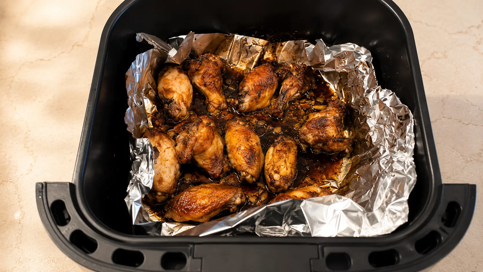 Freshly cooked teriyaki chicken wings in a foil-lined air fryer basket with a glossy caramelized sauce.