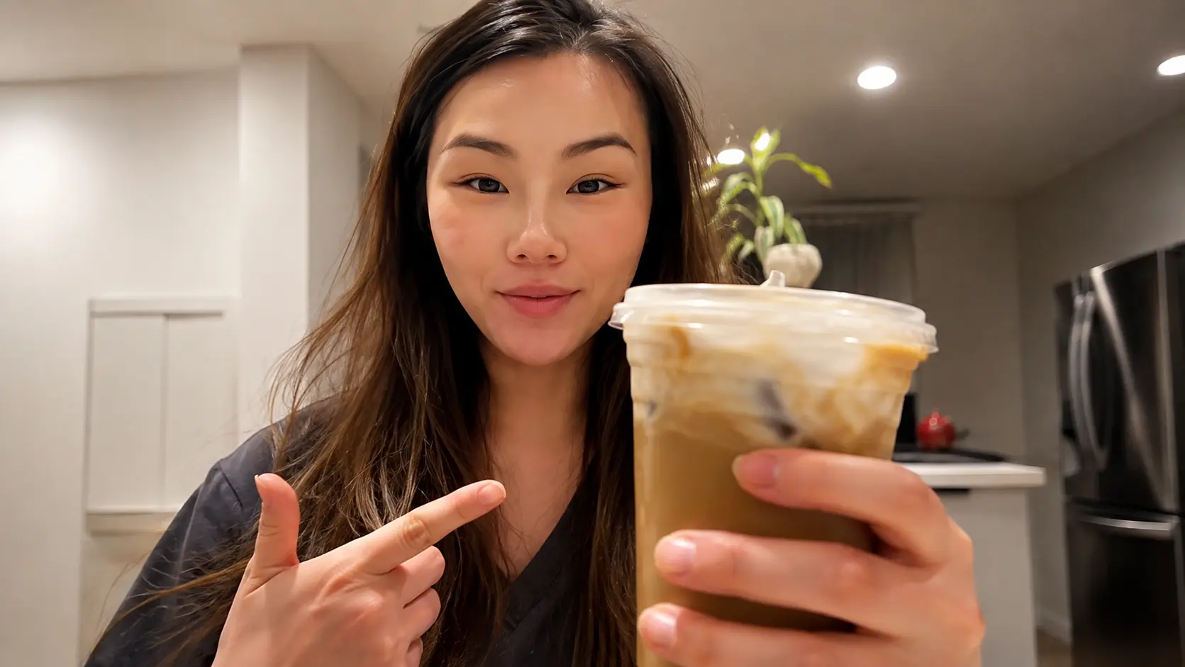 Person holding a large iced coffee toward the camera while pointing at it, standing in a softly lit kitchen with recessed ceiling lights.