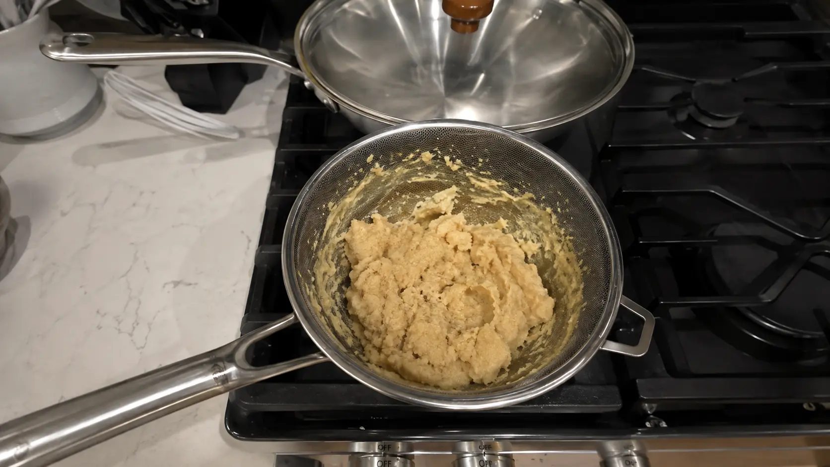 A fine-mesh strainer holds a thick pale dough over a stovetop, with a stainless steel pan and lid in the background.