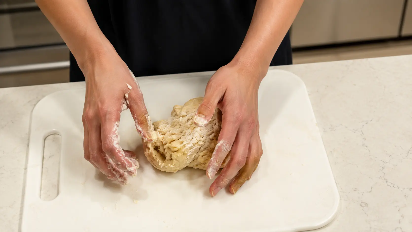 Flour-dusted hands knead a pale dough on a white cutting board set on a light-colored kitchen countertop.
