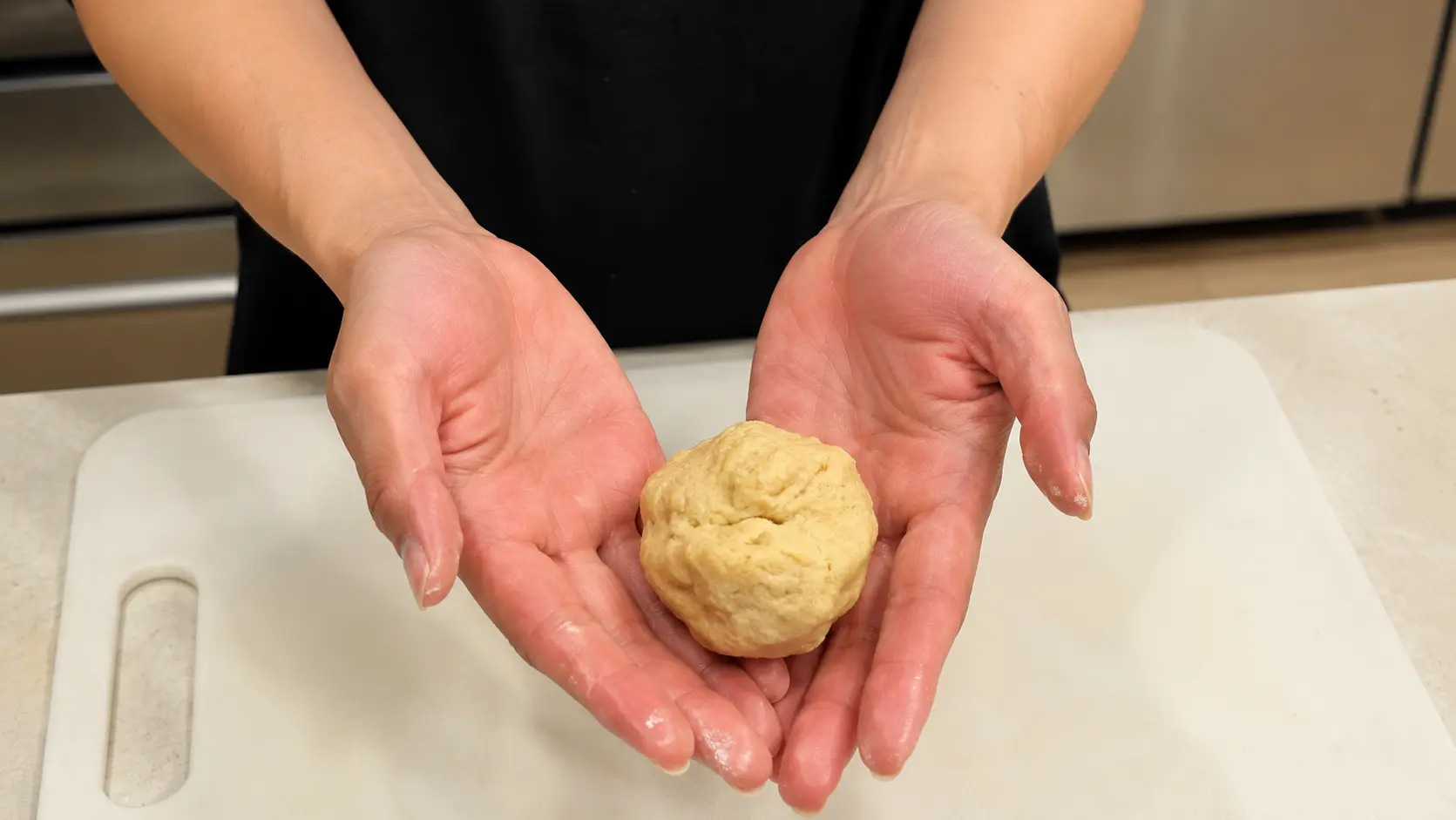 Two hands hold a small round ball of pale dough above a white cutting board on a light-colored kitchen countertop.