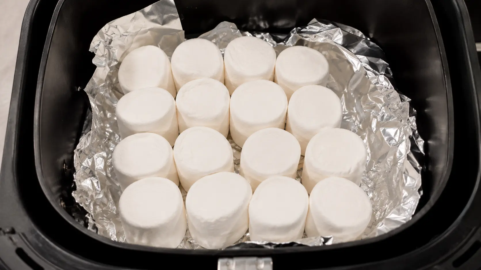 Top-down view of jumbo marshmallows arranged on crumpled aluminum foil inside a black air fryer basket.